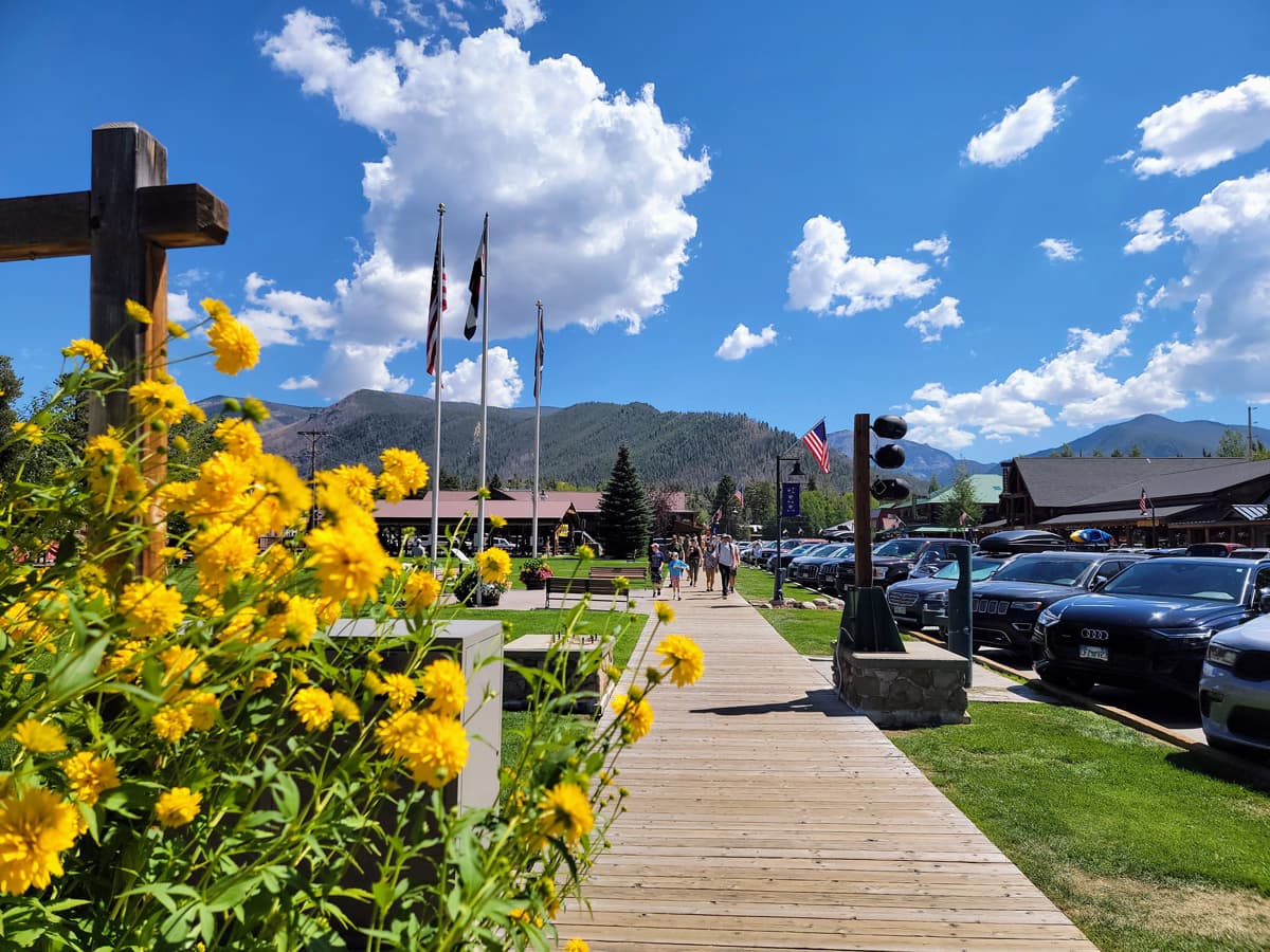 On a blue-sky, summer's day with white clouds, people walk down a historical boardwalk in Grand Lake. Yellow flowers grow in abundance on the left side of the image.