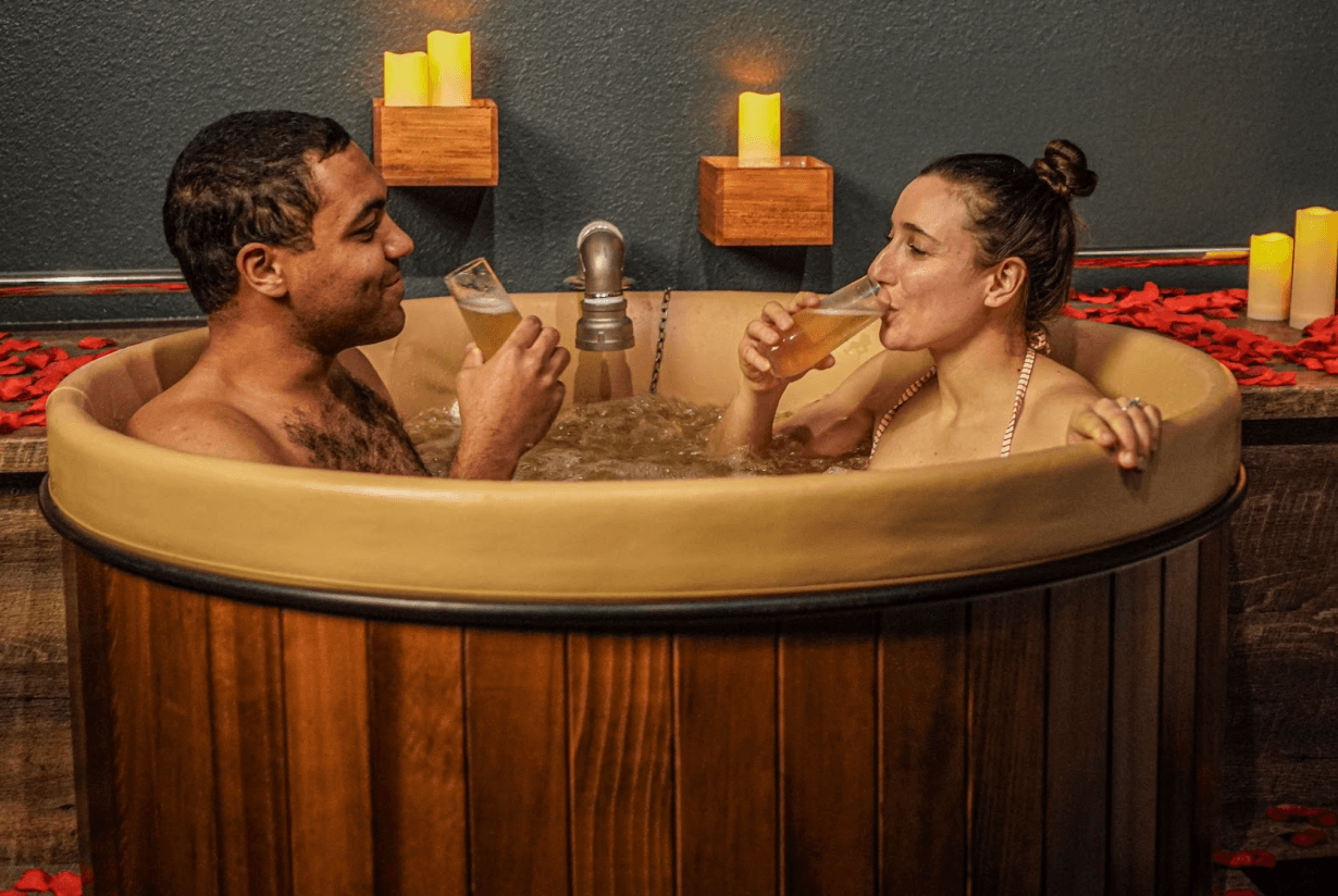 Two people in swimsuits sit in a wooden vat filled with bubbling water and sip tall glasses of pale-yellow beer in Denver, Colorado.