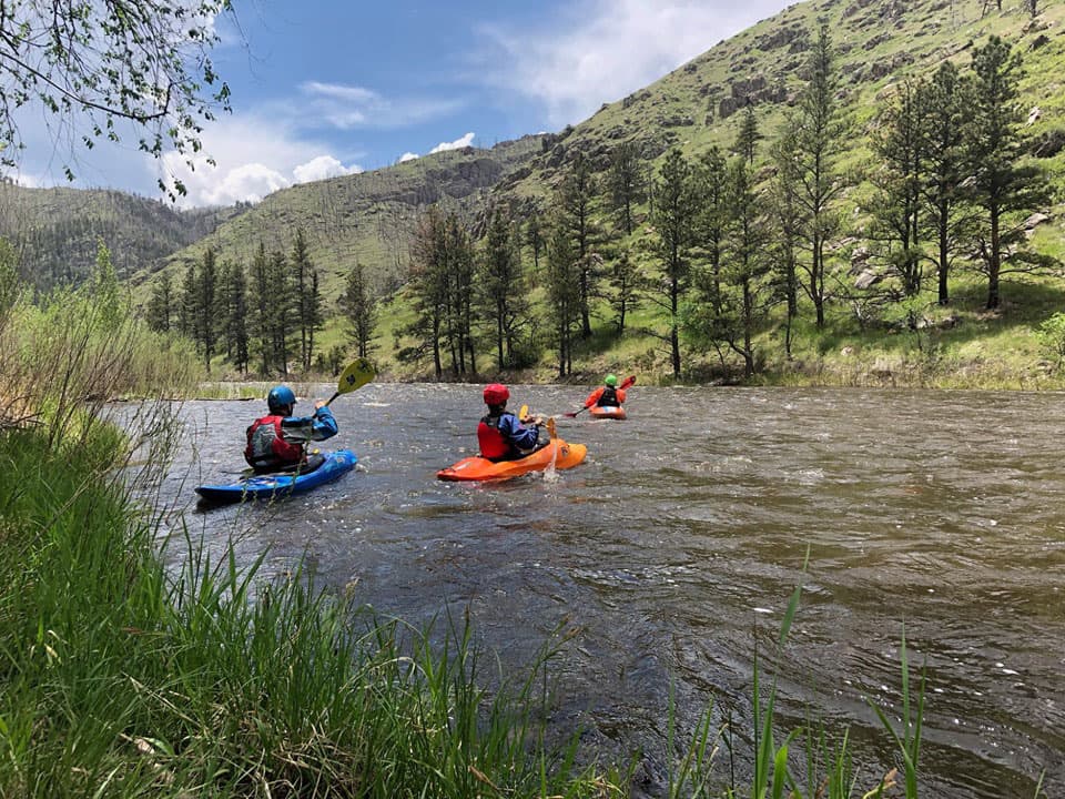 A scenic photo of calm river water running between two grassy areas. There are three student kayakers in the water on blue and orange kayaks wearing protective gear. They are paddling in the water facing away from the camera. In the background are grassy and rocky hills spotted with tall pine trees beneath a mostly blue sky.