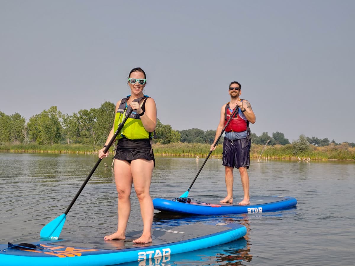 Two happy stand-up paddleboarding students stand up on their blue paddleboards. They are both in swimming attire and are wearing safety vests and sunglasses with paddles in their hands. The water they are on is calm with small rippling waves. In the background is a grassy area with trees and a dusty blue sky.
