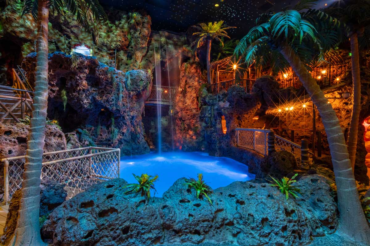 A neon-blue pool is surrounded by fake indoor palm trees and lava with a waterfall flowing into from high above