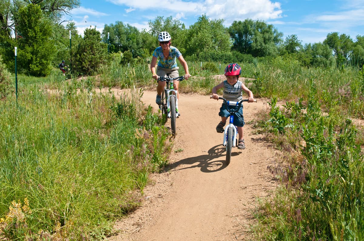 A child and parent ride bikes on a dirt trail surrounded by lush green grass and trees at Valmont Bike Park in Boulder, Colorado