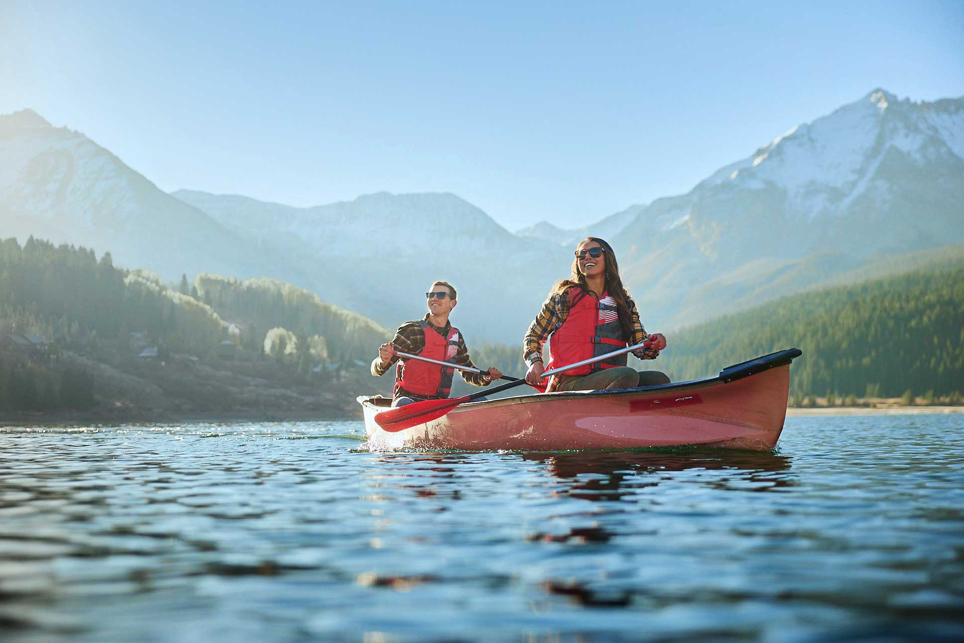 Two people in a red canoe paddle across a lake with mountains behind them