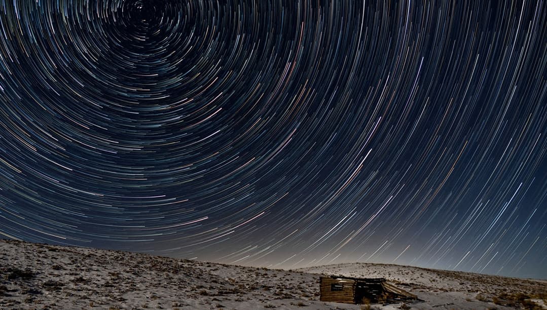 A swirl of star trails at night over an old cabin and snowy ground near Westcliffe, Colorado