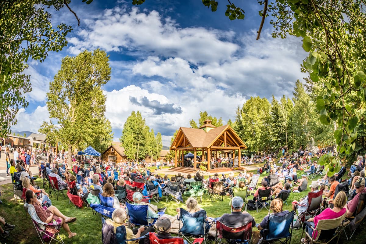 A large group of people sitting outside on a green lawn under a cloud-streaked blue sky listening to a band play in Frisco, Colorado