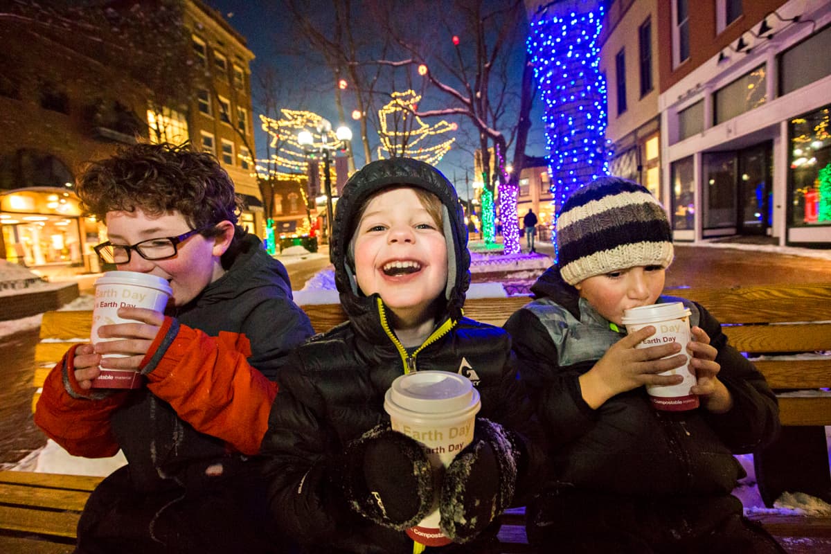 Three children dressed in warm clothing sit on a bench on Pearl Street in Boulder, Colorado. The two kids on the outside are happily sipping a warm drink from to-go cups while the child in the middle is holding onto their cup in mittened hands, staring into the camera with a wide, genuine goofy smile. Behind them are the glimmering holiday lights adorning trees under a winter night sky.