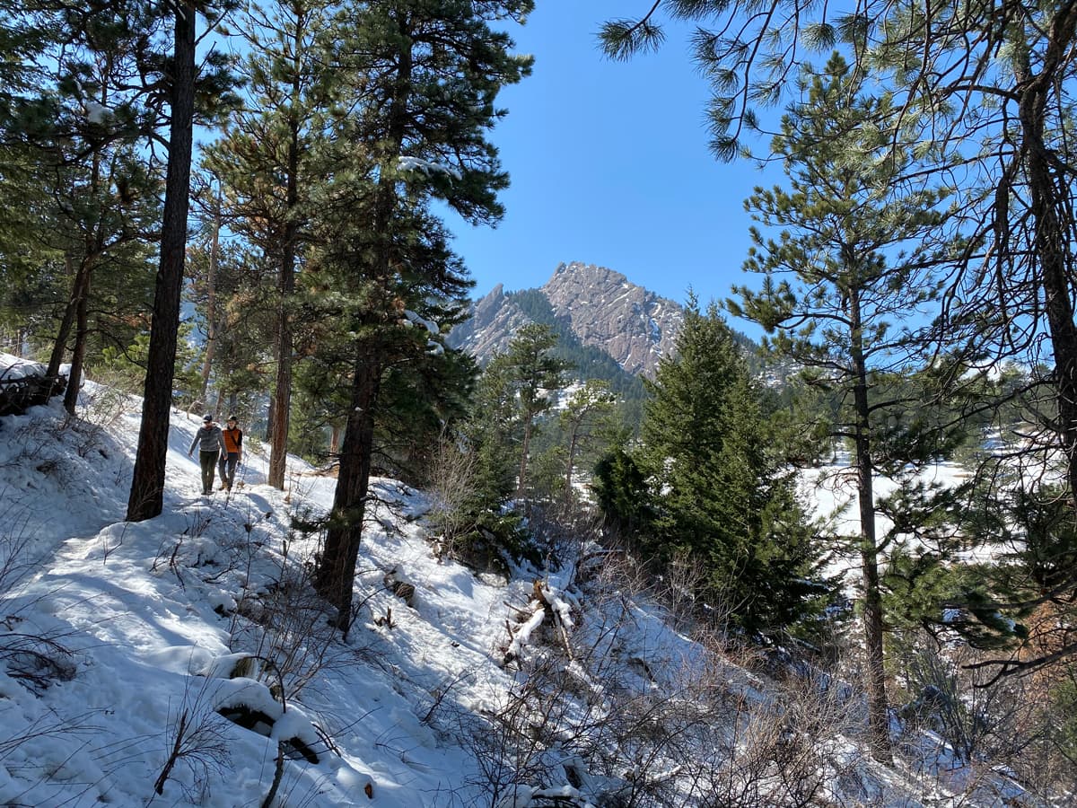 Two hikers to the left side of the photo hike on a snowy trail in Boulder, Colorado. Beside them on an incline is a snow-covered hill peppered with tall pine trees. The sun shines on the snow. In the background is the jagged peak of the Flatirons underneath a dusty blue sky.