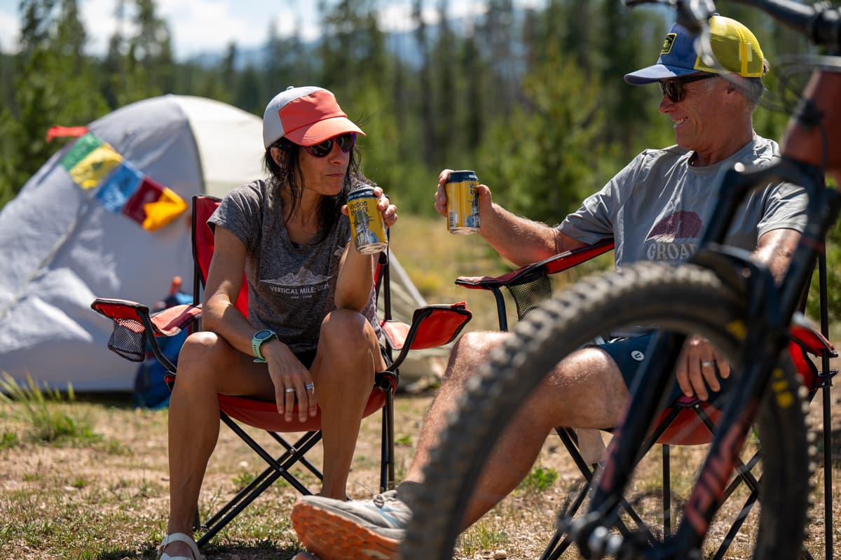 Two people in hats and comfortable outside clothes sit in their camping chairs and cheers cans of beer to each other as the sun shines down on their campsite in Winter Park, Colorado.