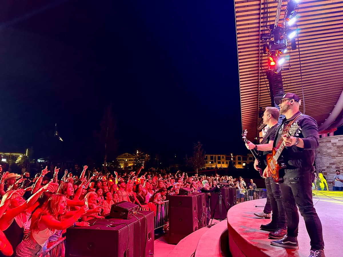 A lively crowd of concert-goers scream and throw their hands in the air as the crowd at a concert in Winter Park. The performers are standing on stage close to the edge, playing guitars.