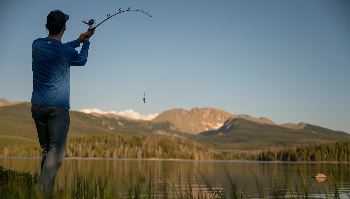 A lone fisher stands in some tall grass lands beside a large body of water in Winter Park, Colorado. Their back is facing the camera as they cast their fly-fishing rod into the lake's rippling surface. The sky above is dusty blue and in the distance, behind the tall, jagged peaks of a mountain, there are puffy white clouds rising up on the horizon.