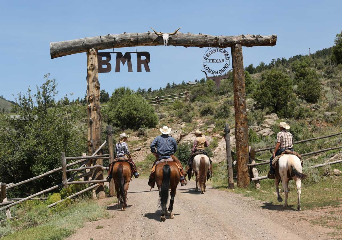 Guests ride horses under the Black Mountain Ranch entrance, made of rough logs