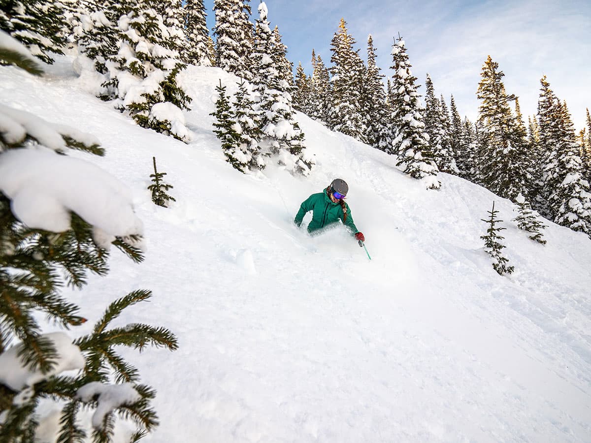 A woman skier in a green jacket waist-deep in snow on a steep mountain surrounded by snowy pine trees