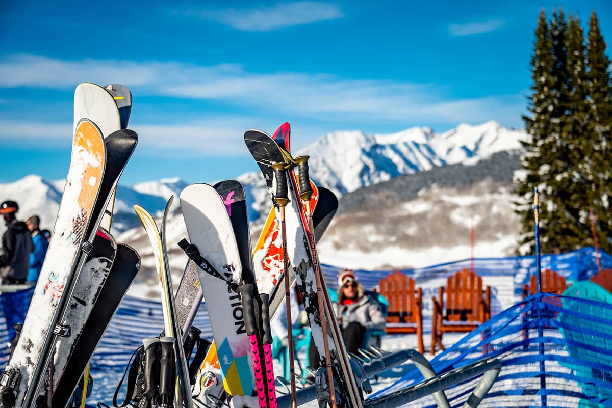 A collection of skis stored on a rack outdoors with a blurred background of mountains in the distance