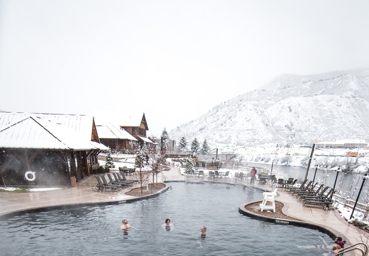People relax in a large, steaming hot springs pool with a backdrop of snow-blanketed mountains at Iron Mountain Hot Springs in Glenwood Springs, Colorado