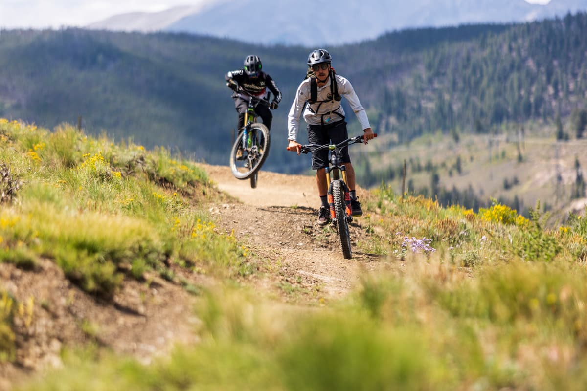 Two bikers race down a mountain singletrack trail and one pops a wheelie on a mountain biking tour with Colorado Adventure Guides