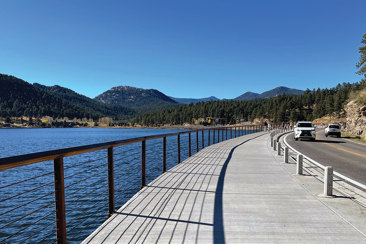 Evergreen Lake Trail with road on the right and the lake on the left, distant mountains in background.