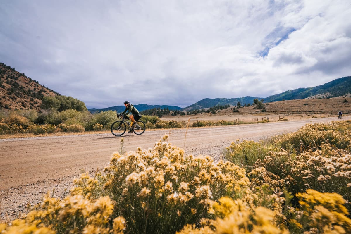 Man gravel biking on a dirt road with mountains behind them and plants off the side of the road.