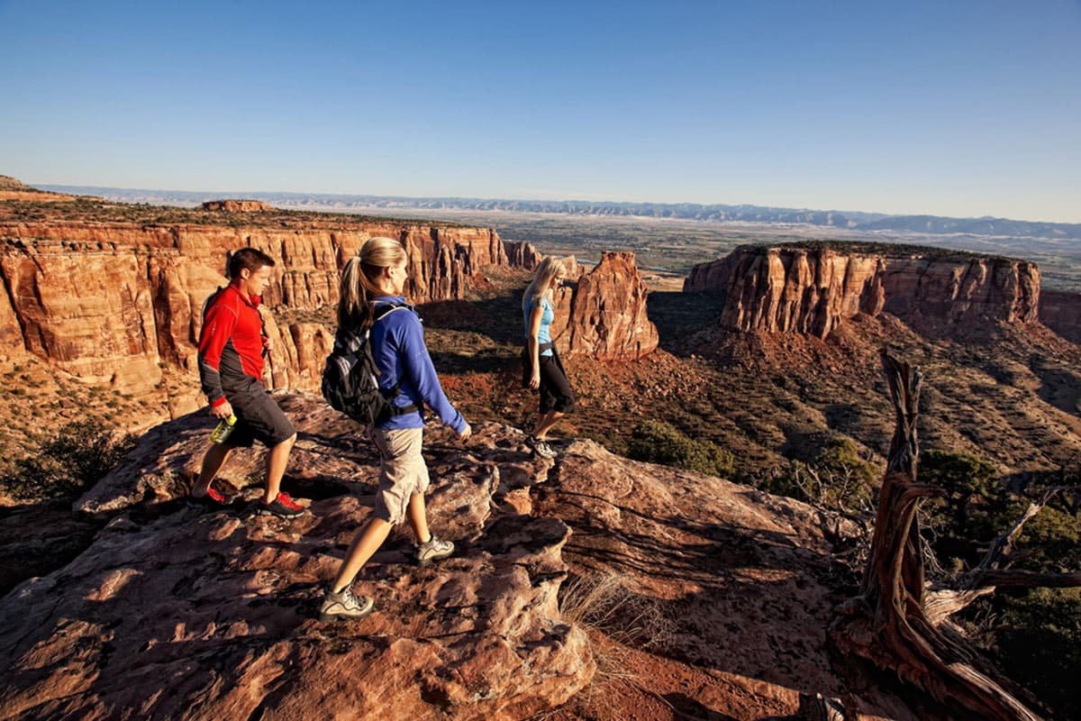Three hikers carefully step on rocks and logs as they make their way to the canyon rim to see the dramatic red-rock cliffs of the monument