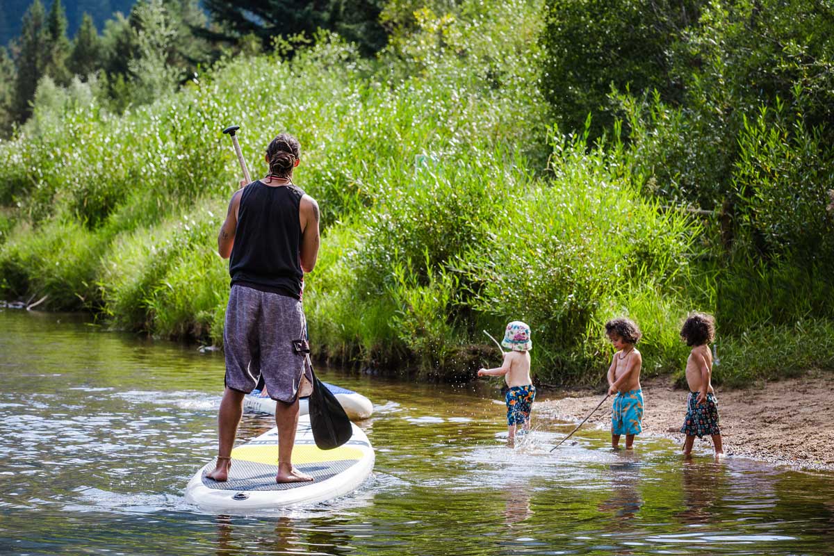 A man steers a yellow stand-up paddleboard toward a river bank where three little kids splash the water with sticks