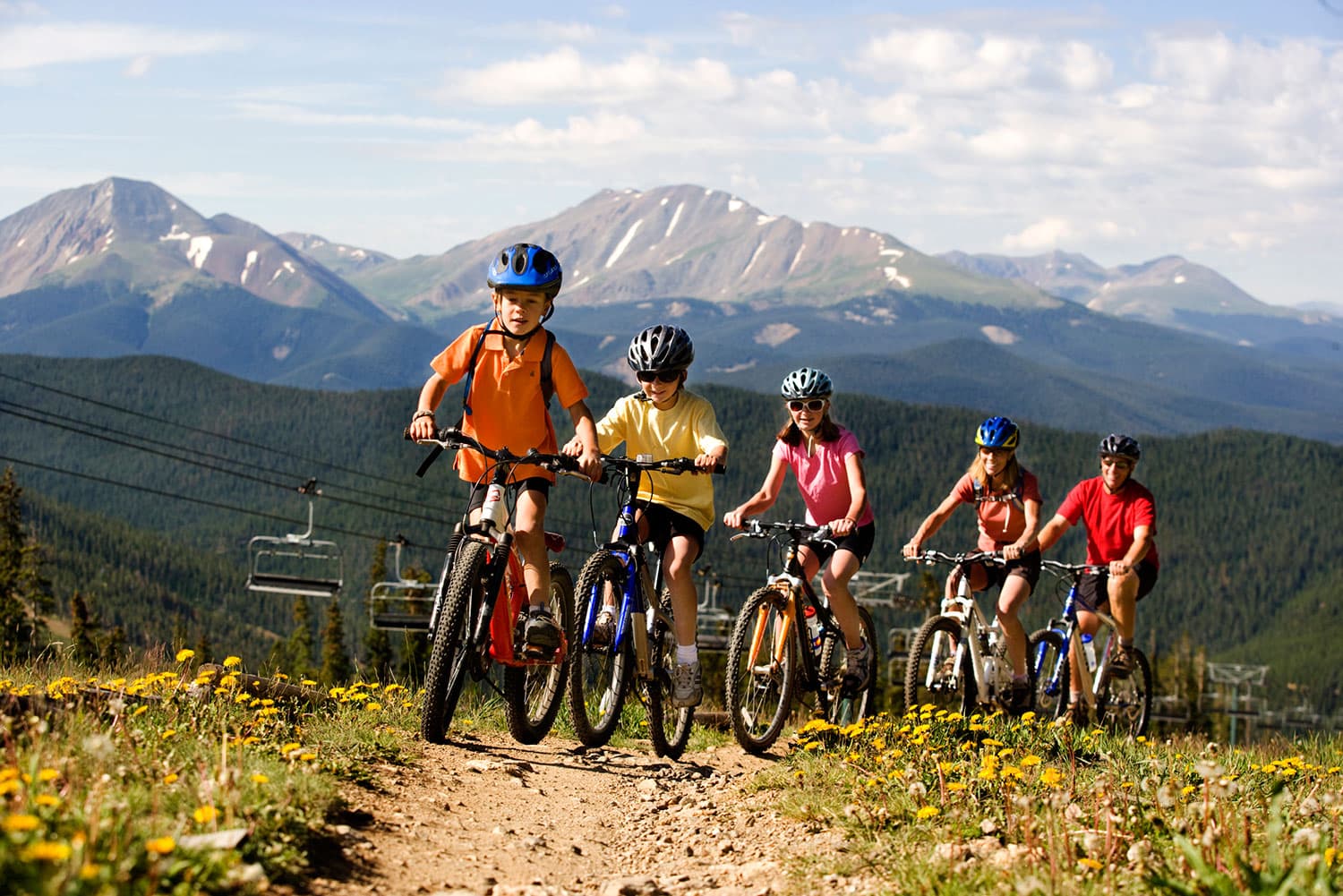 A group of children ride bikes at a ski resort with large mountains surrounding them