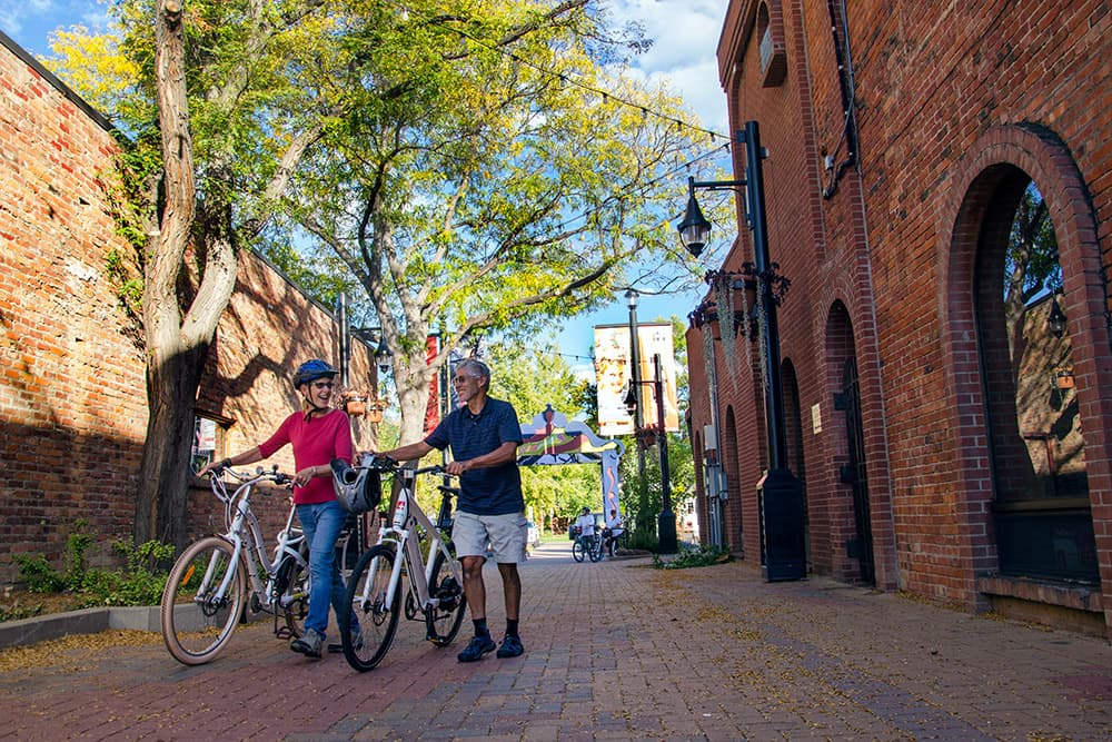 A couple walks their bikes through a brick street with brick buildings on both sides. Up above green-leafed trees lead to a blue sky.