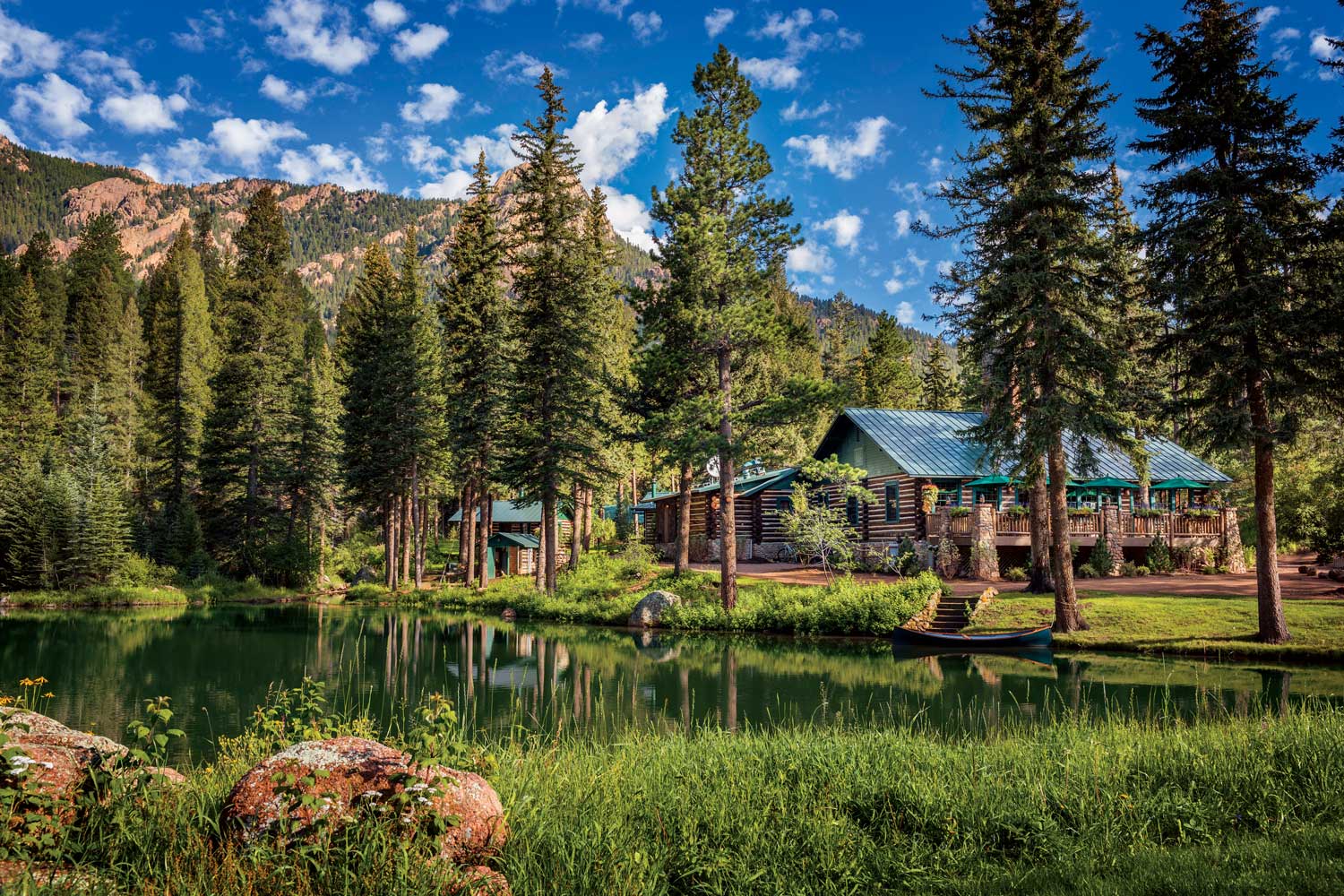 A cozy wood cabin with a faded-blue roof is set next to a pine tree-lined pond in Colorado. A rugged mountain face watches over the area in the background.