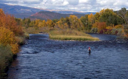 A fisherman stands in the middle of a dark blue, wide river. On both riverbanks the trees are red, yellow and dark green with leaves changing in the fall. In the background a blue sky covered in white clouds sits above a mountain.