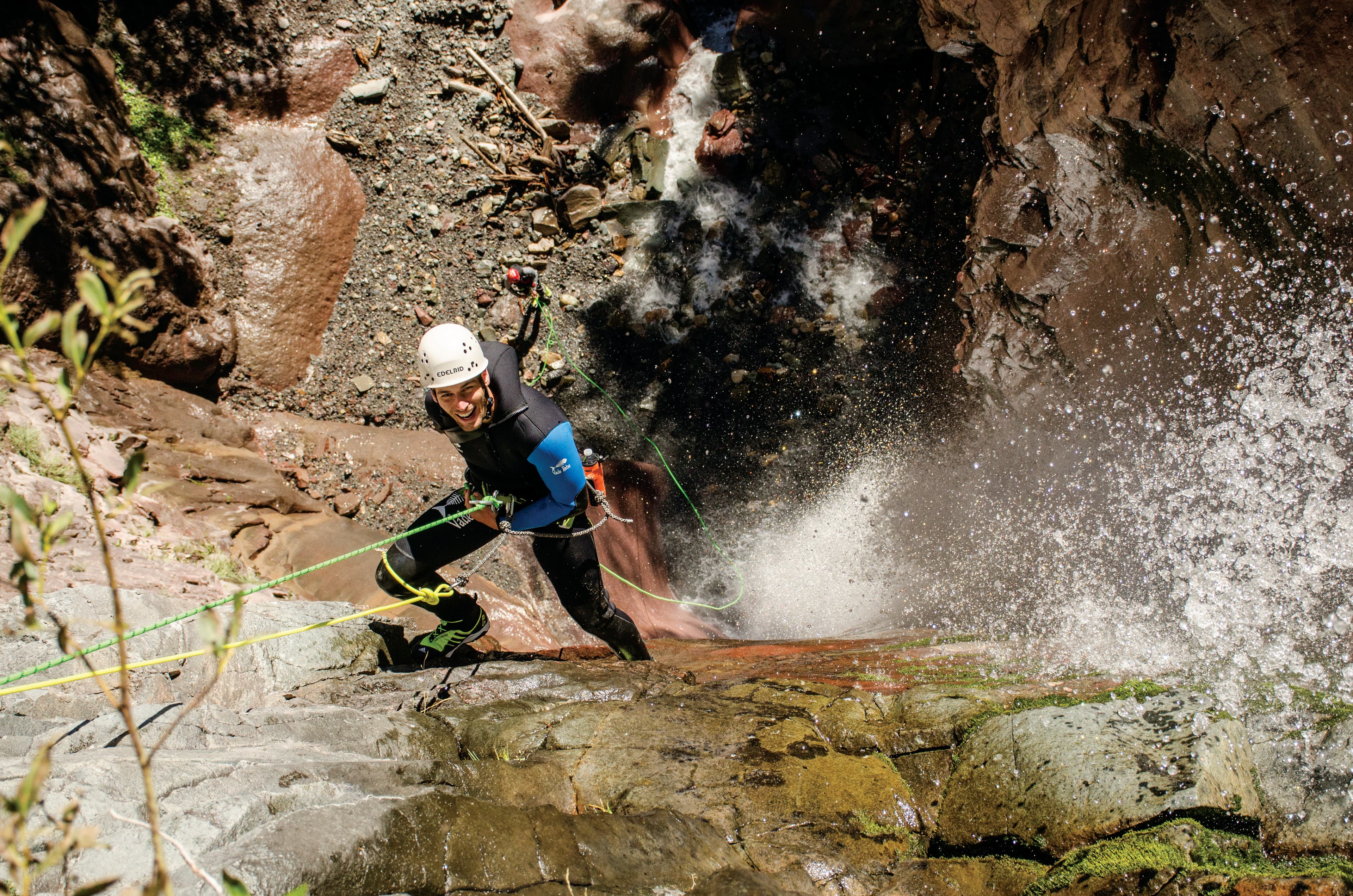 A climber in a wet suit, helmet and climbing harness smiles and looks up as they scale the rocky wall next to a flowing waterfall in Colorado.