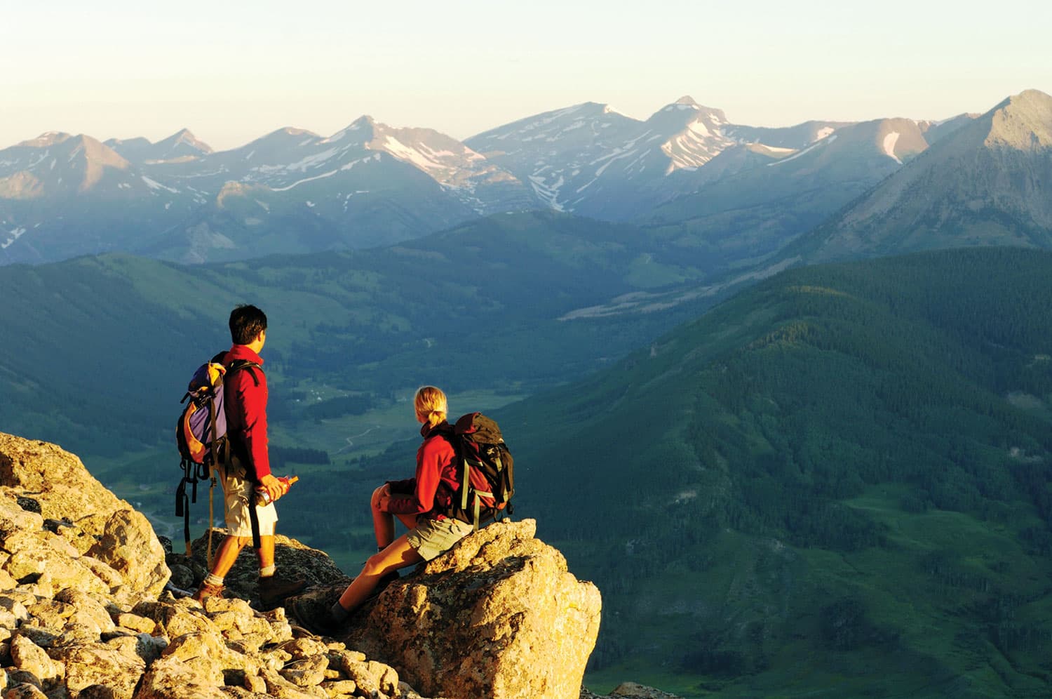 Two hikers in matching red jackets take a break at a rock outcrop near Crested Butte Mountain Resort in Colorado. They admire the view of the verdant valley and majestic peaks.
