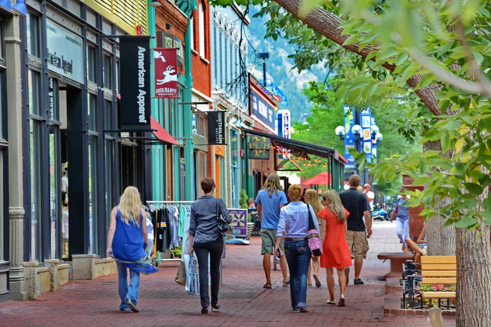 Groups of shoppers walk down the brick pedestrian paths of Pearl Street Mall in Colorado. Some hold shopping bags while others look at the colorful storefront along the street.