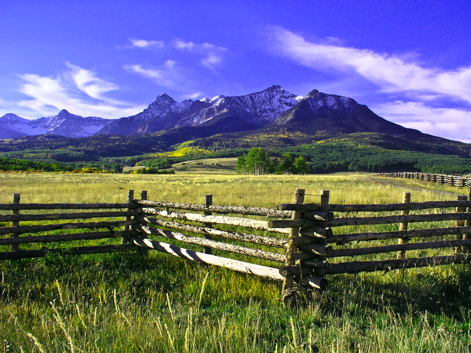 A wooden fence zigzags through a meadow with craggy peaks in the background