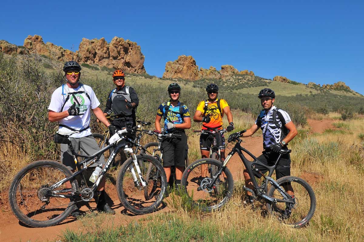Five mountain bikers wearing helmets pose for a photo on a dirt track in Loveland, Colorado. Tall grasses surround the track on either side and craggy rock formation appear in the background.
