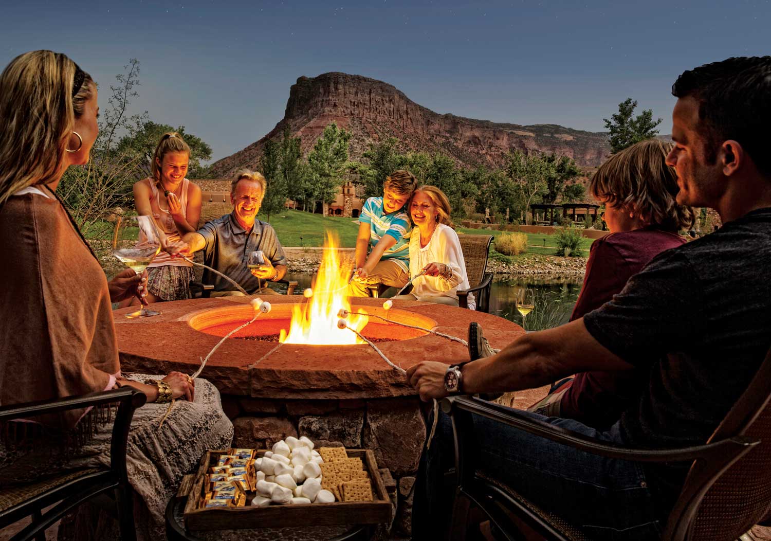 A multi-generational family gathers around a wide, stone-lined fire pit at Gateway Canyon Resort in Colorado, and roasts marshmallows over the fire.