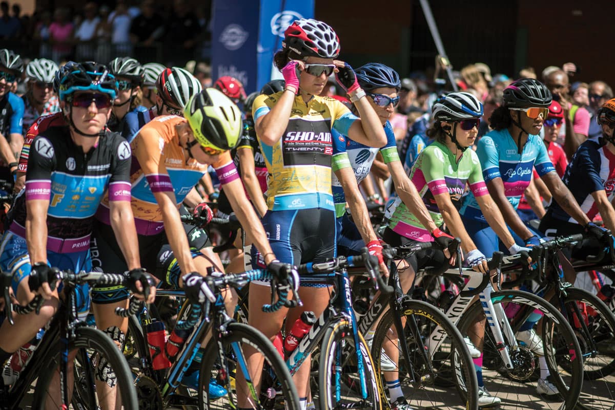 A cyclist stands and straddles their bike to adjust their sunglasses before the start of a race in Colorado featuring dozens of riders. They are side-by-side in a line at the front of the group with six other athletes.