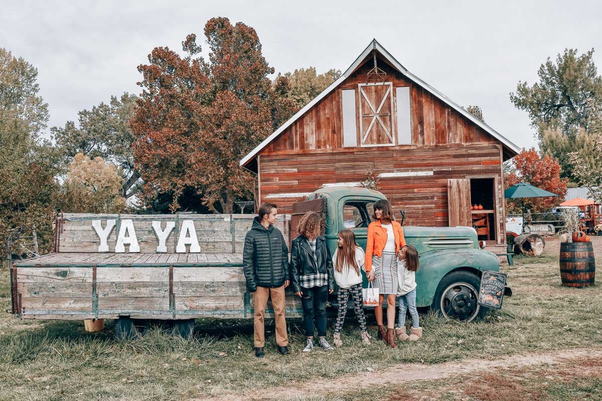 A family poses with an old apple picking truck that has large white YA YA letters. In the background a wooden building sits in a grove of trees.