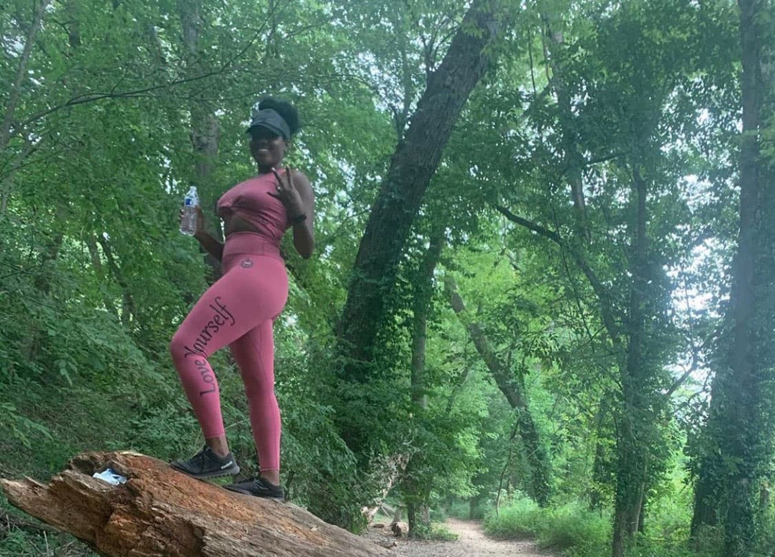 A hiker poses on a fallen tree trunk in the middle of the Colorado woods. They wear pinkish-red activewear and hold a bottle of water in their hand.