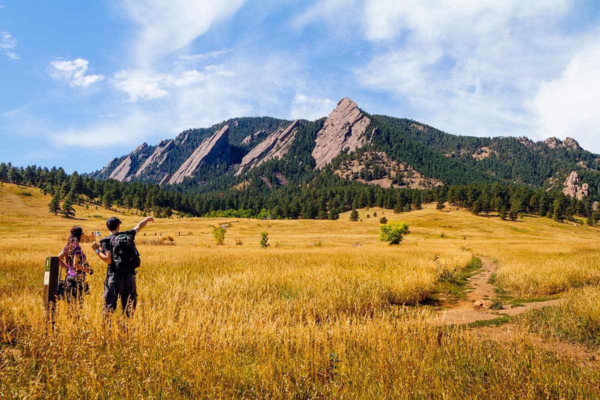 Tall prairie grasses shine a deep gold beneath the late-summer sun. Several dirt paths wind through the grasses. Two hikers discuss the path leading to the tall, rocky peaks in the distance.