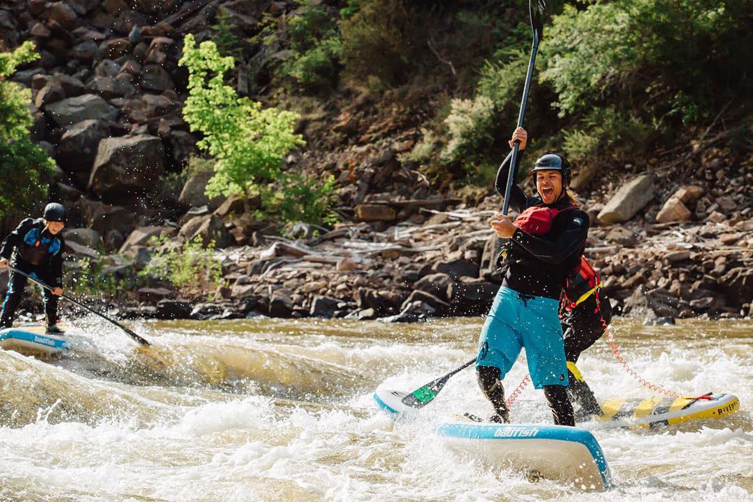 People in helmets laugh as they balance on a rapid on a stand-up paddleboard in the river