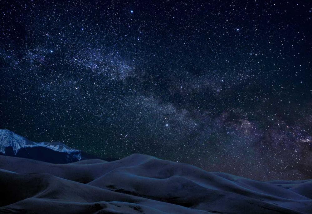 The night sky with stars decorating it sits above the Great Sand Dunes. In the distance snow-covered peaks sit.