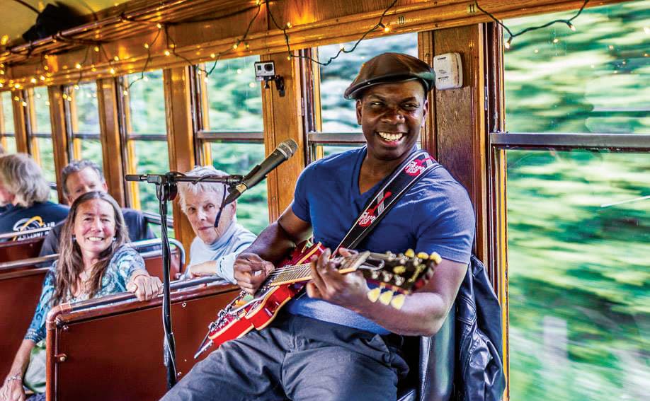A musician smiles as they play a stop sign-red, electric guitar and sing to smiling passengers in a car of the Durango & Silverton Narrow Gauge Railroad’s Blues Train.