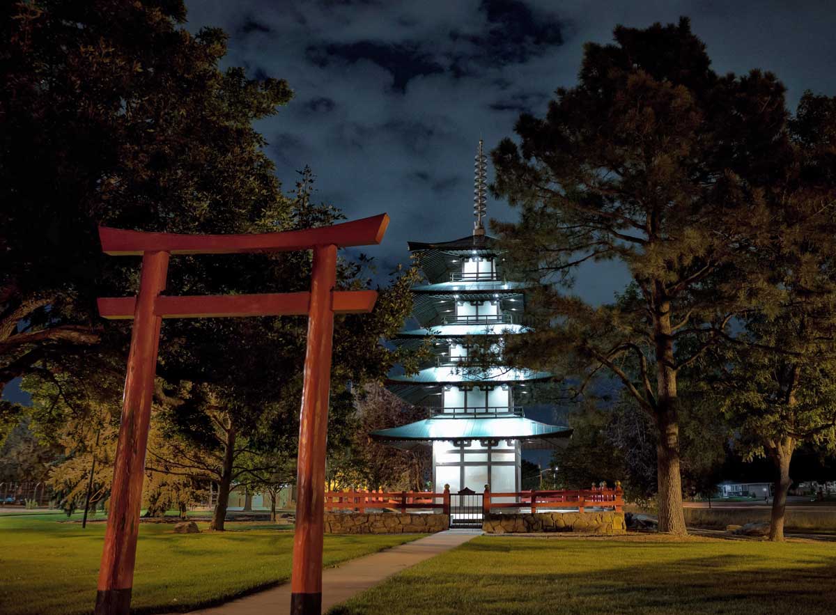 A red gate on the left side of the image welcomes people to the five-layered Tower of Compassion in Kanemoto Park at night. It's surrounded by trees and green grass.
