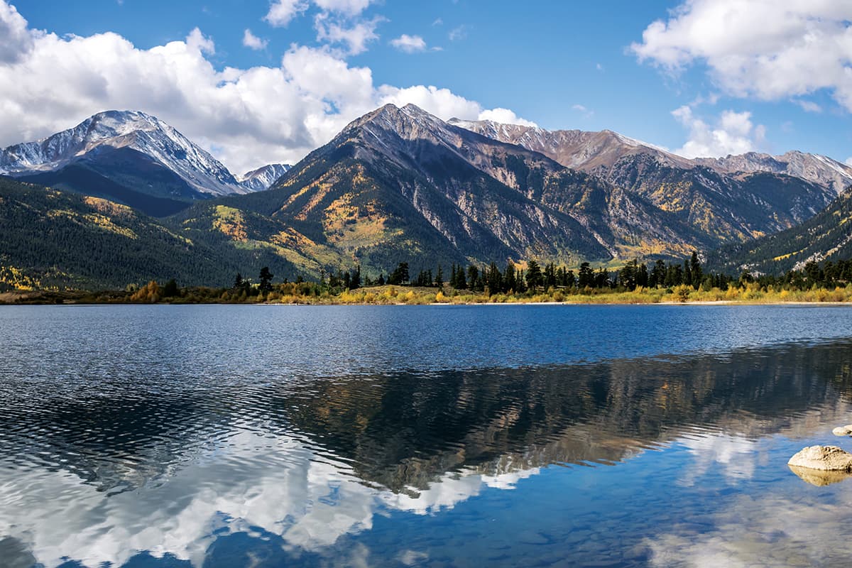 A lake reflects the image of a mountain range dusted with snow at the peaks and bursts of gold and orange fall foliage on an otherwise evergreen mountainside in Colorado.