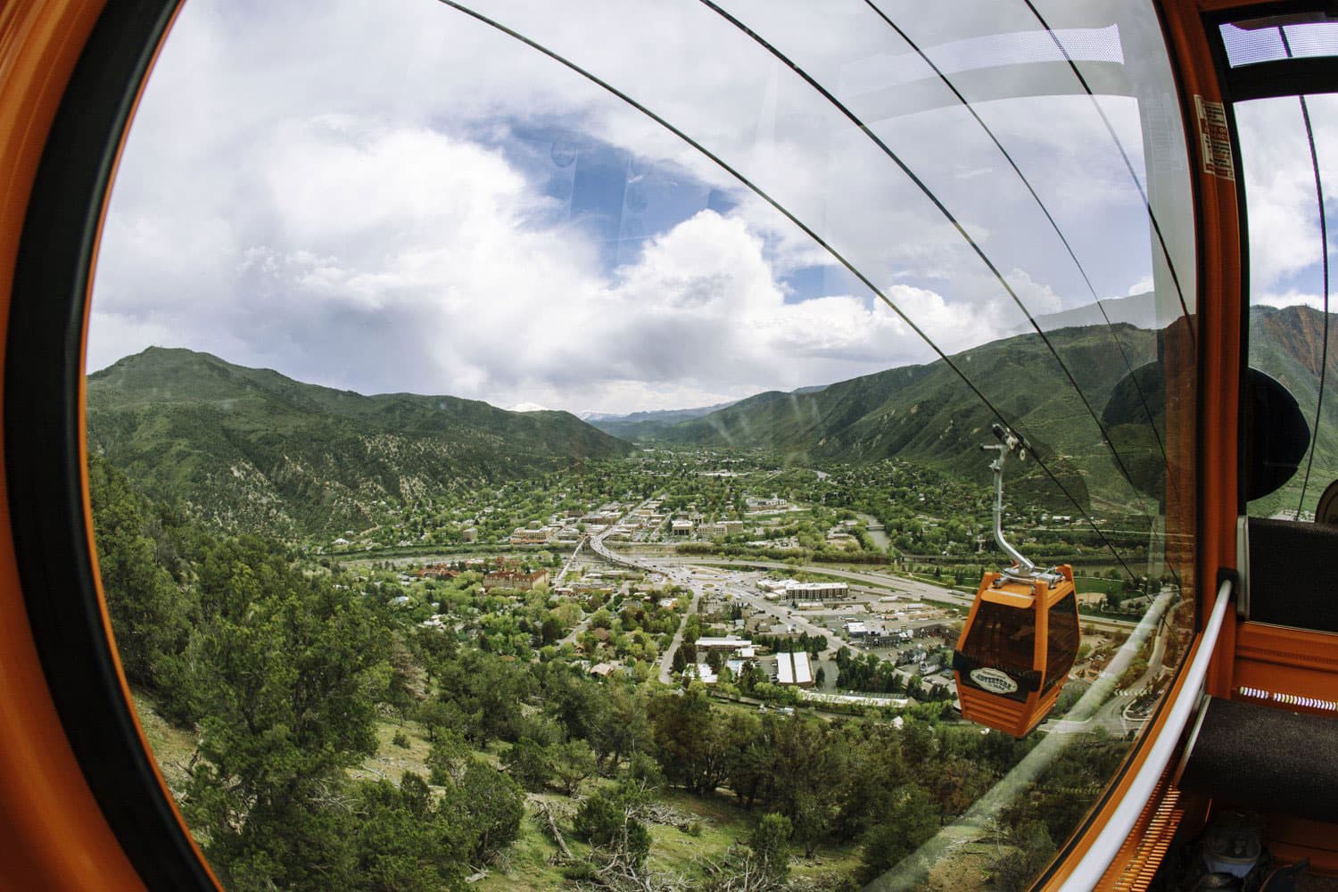 The view from a Glenwood Gondola shows the town laid out between lush, verdant mountain ridges and an orange gondola traveling down the cables.