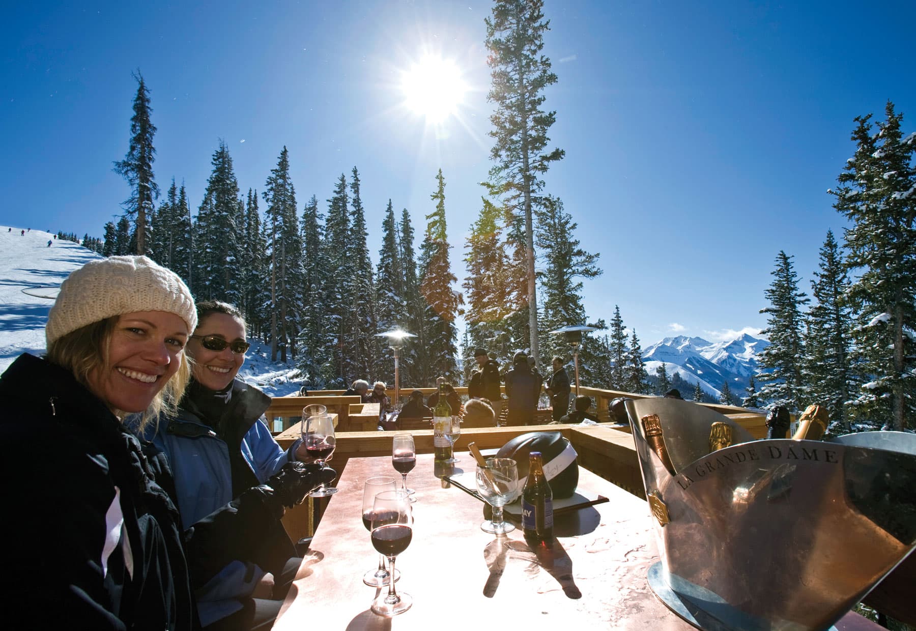 People smiling with glasses of red wine with a ski mountain and trees in the background at Alpino Vino in Telluride, Colorado