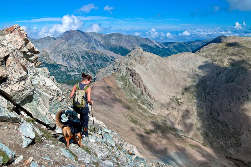 A tan and black dog follows behind a person wearing a green backpack. Together they traverse the rocky landscape of a mountainside in Colorado.