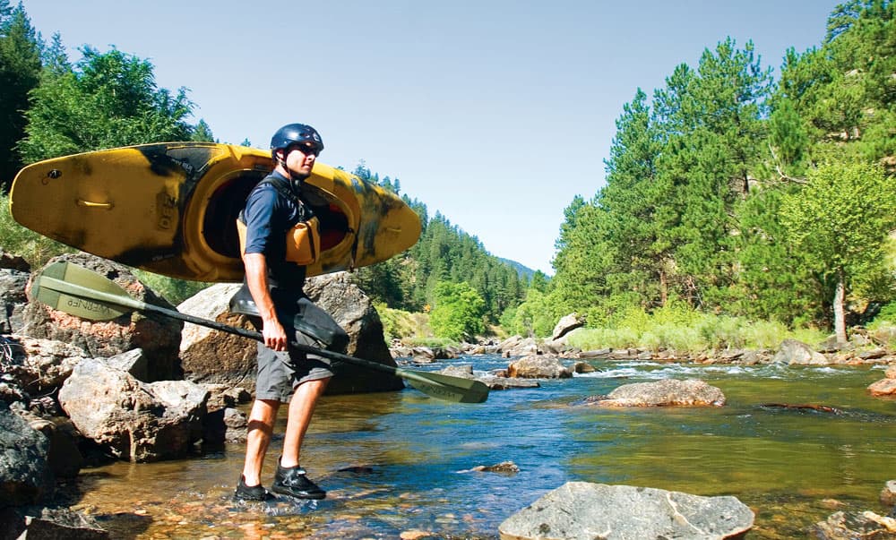 A kayaker hoists their kayak over their shoulder with one hand and holds their paddle in the other hand. They walk along the rocky shallow banks of the Cache la Poudre near Fort Collins.