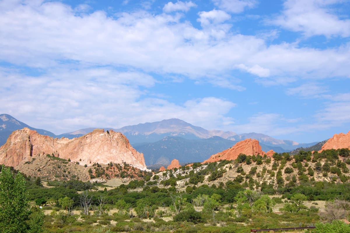 Rolling foothills with low shrubs and trees lead up to the red rock ridges of the Garden of the Gods. Two rows of mountain ranges loom in the distance.