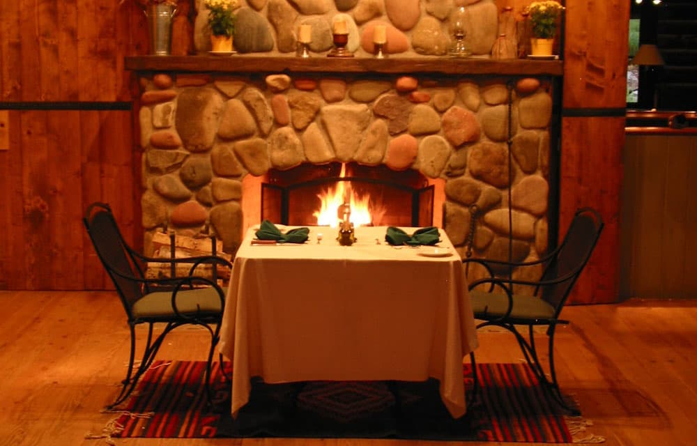 A vibrant fire blazes in the hearth of a red and gray stone fireplace in Crawford, Colorado. Before the fireplace is a cozy table set for two.
