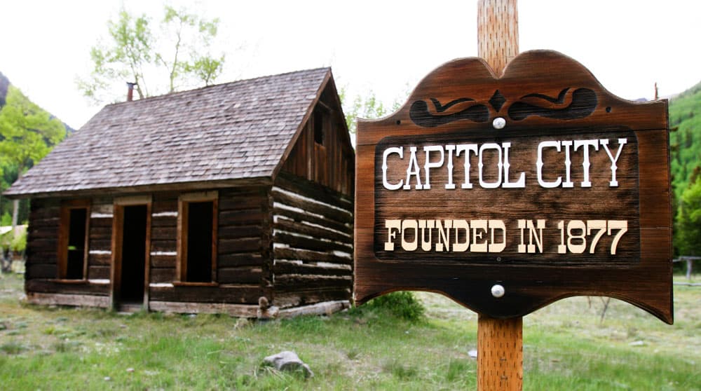 A wooden sign says Capitol City with a long cabin in the background and green grass on the ground.