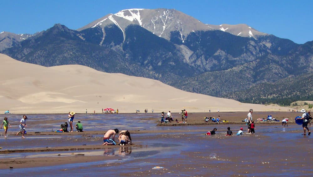 Groups of children and adults frolic and swim about in the shallow waters of the Medano Creek which lies at the base of great sand dunes and mountains in Colorado.
